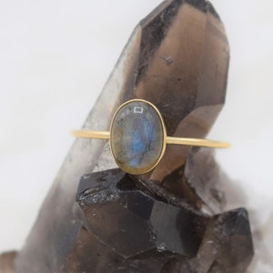 Puede incluir: Un anillo de oro con una piedra labradorita ovalada. La piedra preciosa tiene tonos azules y grises. El anillo tiene una fina banda de oro. El anillo se muestra sobre una superficie oscura y texturizada.