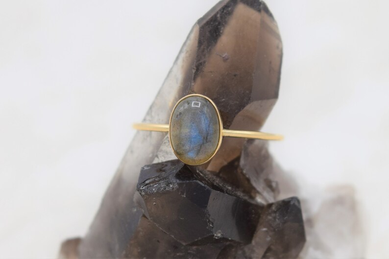 Puede incluir: Un anillo de oro con una piedra labradorita ovalada. La piedra preciosa tiene tonos azules y grises. El anillo tiene una fina banda de oro. El anillo se muestra sobre una superficie oscura y texturizada.
