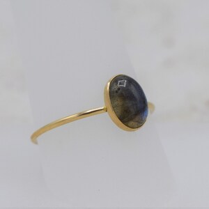 Puede incluir: Un anillo de oro con una piedra labradorita ovalada e iridiscente. La piedra muestra una mezcla de tonos azules, grises y amarillos. El anillo tiene una banda fina y delicada.