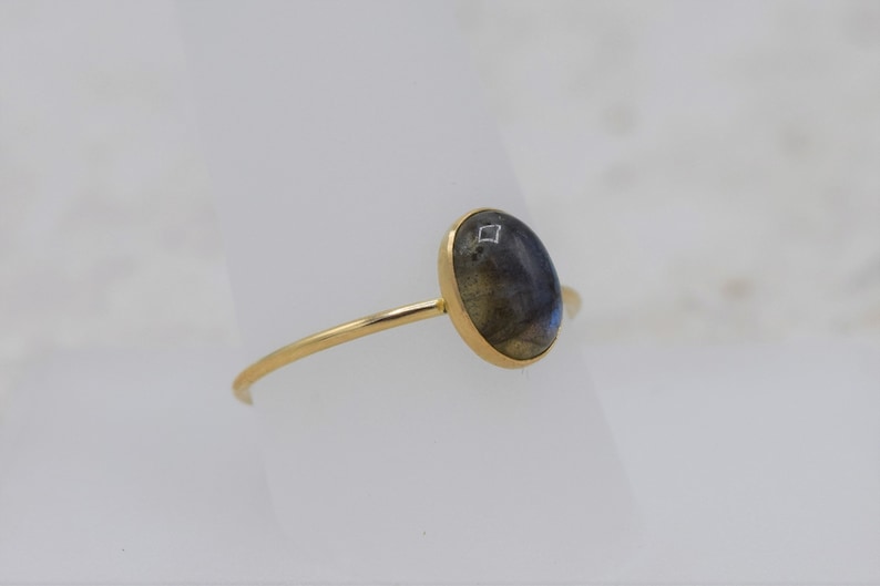 Puede incluir: Un anillo de oro con una piedra labradorita ovalada e iridiscente. La piedra muestra una mezcla de tonos azules, grises y amarillos. El anillo tiene una banda fina y delicada.