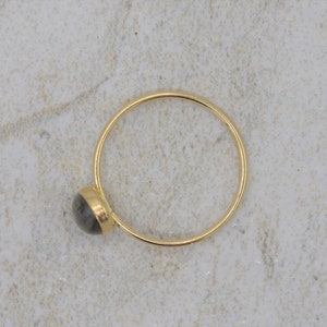 Puede incluir: Un anillo de oro con una peque&ntilde;a piedra preciosa redonda y gris. La banda del anillo es fina y circular, y la piedra preciosa est&aacute; engastada en un bisel de oro. El anillo se muestra sobre una superficie clara y texturizada.