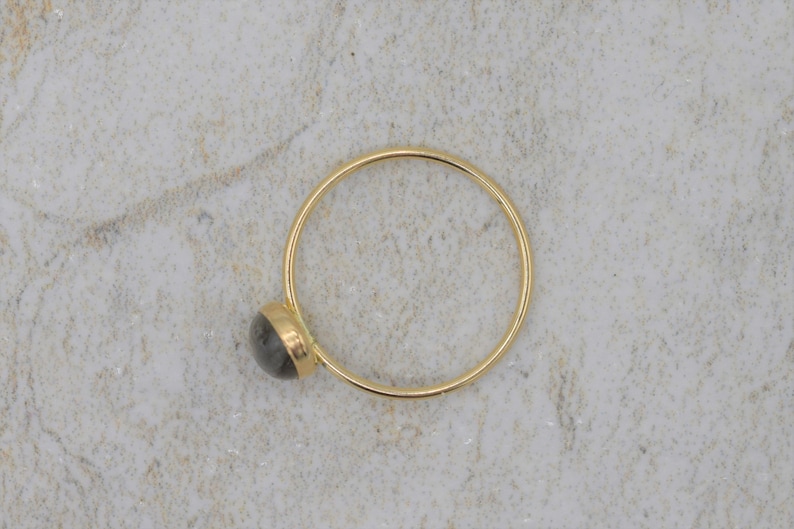 Puede incluir: Un anillo de oro con una peque&ntilde;a piedra preciosa redonda y gris. La banda del anillo es fina y circular, y la piedra preciosa est&aacute; engastada en un bisel de oro. El anillo se muestra sobre una superficie clara y texturizada.