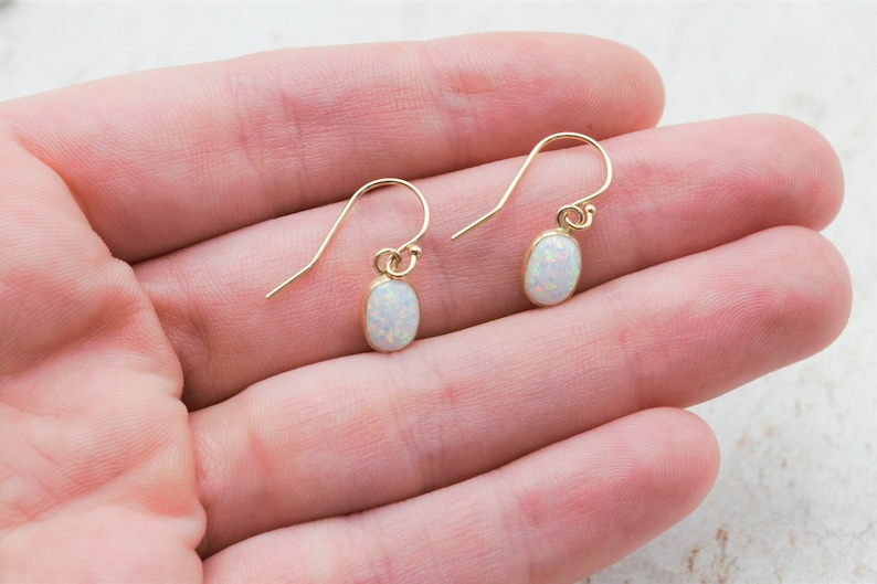May include: A pair of gold earrings with white opal stones. The earrings are oval shaped and dangle from a simple hook.
