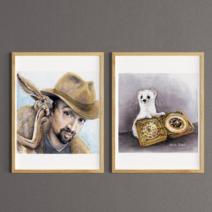 Puede incluir: Dos impresiones artísticas enmarcadas. Una presenta a un hombre con sombrero y un conejo, la otra a una comadreja blanca con un reloj dorado. Ambas son acuarelas con marcos de madera, expuestas contra una pared gris.