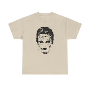 May include: A beige t-shirt with a black and white illustration of a woman's face with tattoos. The woman has dark hair and is wearing a necklace.