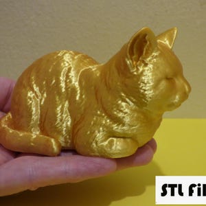 May include: A gold-colored 3D printed cat figurine. The cat is lying down with its head resting on its paws. The figurine has a smooth, shiny finish.