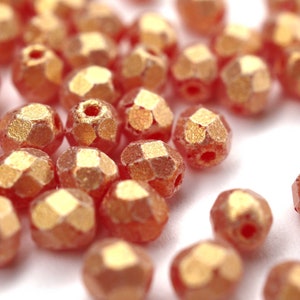 Op de afbeelding: Een close-up van een stapel kleine, gefacetteerde, iriserende kralen. De kralen zijn licht perzikkleurig met een gouden metallic glans.
