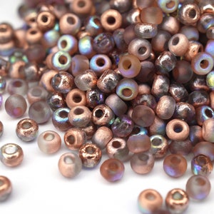 Könnte beinhalten: Ein Haufen kleiner, runder, irisierender Glasperlen in Kupfer- und Brauntönen. Die Perlen haben eine matte Oberfläche und sind auf einer weißen Oberfläche verstreut.