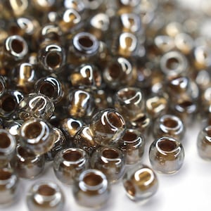 May include: A pile of small, round, iridescent glass beads in shades of brown and gold. The beads have a metallic sheen and are scattered on a white surface.