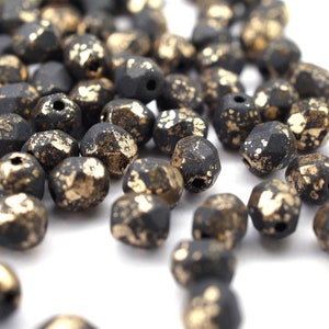 Könnte beinhalten: Eine Streuung von schwarzen facettierten Glasperlen mit goldenen Flecken. Die Perlen haben eine raue, strukturierte Oberfläche.
