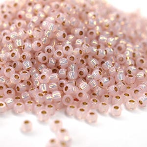 Könnte beinhalten: Ein großer Haufen kleiner, runder, rosa Glasperlen mit goldener Metallic-Oberfläche. Die Perlen sind undurchsichtig und haben einen leichten irisierenden Schimmer.