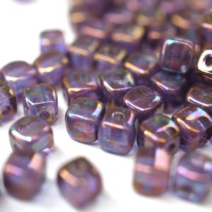 Könnte beinhalten: Nahaufnahme eines Haufens irisierender, lila Würfelperlen. Die Perlen haben einen metallischen Glanz mit Gold- und Blaunuancen. Jede Perle hat ein Loch zum Auffädeln. Ideal für Schmuckherstellung und Bastelarbeiten.