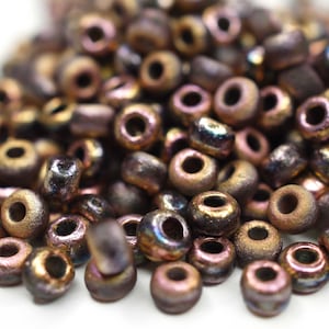 Op de afbeelding: Een hoop kleine, ronde, iriserende kralen in tinten bruin en goud. De kralen hebben een metallic glans en zijn verspreid over een wit oppervlak.