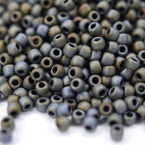 Könnte beinhalten: Ein Haufen kleiner, runder, grauer und brauner Perlen. Die Perlen haben eine matte Oberfläche und sind auf einer weißen Oberfläche verstreut.