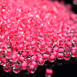 Könnte beinhalten: Ein Haufen kleiner, klarer Glasperlen mit einer rosa irisierenden Beschichtung. Die Perlen sind rund und haben ein Loch in der Mitte.