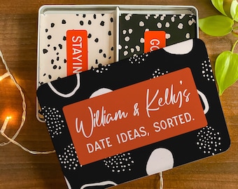 May include: A black and white tin box with a red and white label that says "William & Kelly's DATE IDEAS, SORTED." The tin box has a black and white polka dot pattern and white abstract shapes. The tin box contains two sets of cards with black and white polka dot patterns.