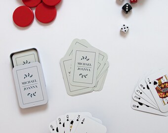 May include: A set of playing cards with a black and red design, a tin box with a white card inside that says "Michael Joanna" and a stack of white cards with the same text. There are also red and white dice and a white number 64.