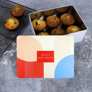 May include: A white metal tin with a red, white and blue geometric design. The lid of the tin has the text "Millie's Sweet Treats" printed on it. The tin is open and filled with muffins.