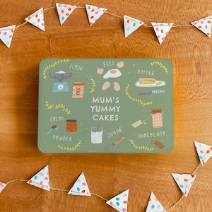 May include: A green rectangular baking tin with illustrations of cake ingredients, including flour, eggs, butter, cocoa powder, sugar, and chocolate. The tin has the text "Mum's Yummy Cakes" in the centre.