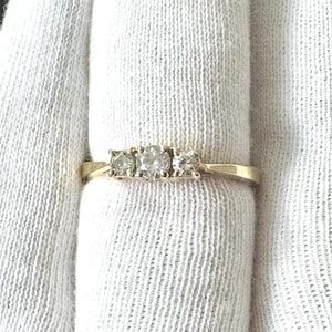 May include: A gold ring with three clear, round diamonds set in a row. The ring is displayed on a white fabric background. The diamonds are of varying sizes, with the center stone being the largest.