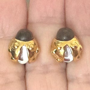 May include: A pair of gold-toned ladybug stud earrings. Each earring features a dark brown dome and a gold-colored body with white and burgundy accents. The earrings are designed with a textured surface.