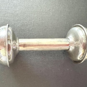 May include: A polished silver-toned baby rattle featuring a smooth cylindrical handle connected to two rounded, dome-shaped ends. The metallic surface reflects light, creating a bright, shiny appearance across the entire object.