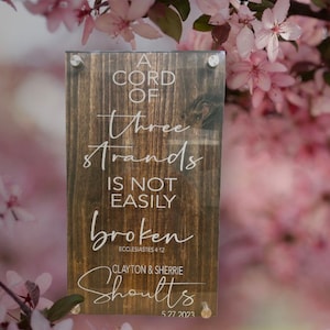 Op de afbeelding: Een houten bord met een witte inscriptie die luidt "A cord of three strands is not easily broken, Ecclesiastes 4:12". Onder de inscriptie staat de tekst "Clayton & Sherrie Shoults, 5.27.2023".