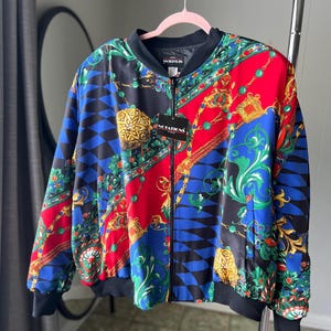 May include: A black bomber jacket with a colorful, patterned design featuring gold chains, red, blue, and green accents. The jacket has a zipper closure and a ribbed collar.
