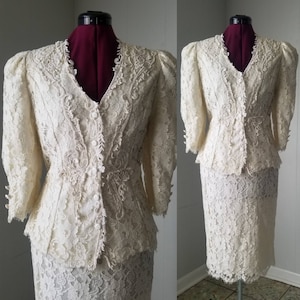 May include: A white lace dress suit with a fitted jacket and a skirt. The jacket has a V-neckline and a button-up front. The skirt is knee-length and has a scalloped hem.
