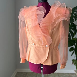 May include: A peach-colored blouse with a ruffled neckline and sheer sleeves. The blouse has a double-breasted closure with two buttons.