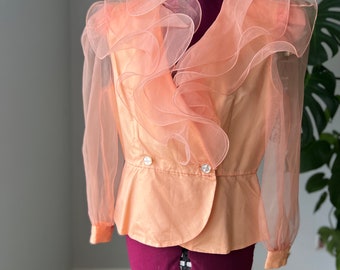 May include: A peach-colored blouse with a ruffled neckline and sheer sleeves. The blouse has a double-breasted closure with two buttons.