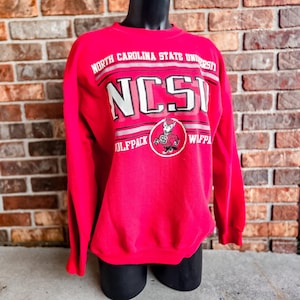 K&ouml;nnte beinhalten: Ein leuchtend rotes Langarm-Sweatshirt mit Rundhalsausschnitt, das den Text NORTH CAROLINA STATE UNIVERSITY und NCSU in wei&szlig;er und schwarzer Schrift zeigt. Eine kreisf&ouml;rmige Grafik des Wolfpack-Maskottchens ist mittig unter dem Text platziert.