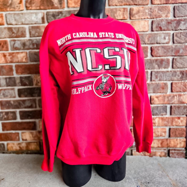 K&ouml;nnte beinhalten: Ein leuchtend rotes Langarm-Sweatshirt mit Rundhalsausschnitt, das den Text NORTH CAROLINA STATE UNIVERSITY und NCSU in wei&szlig;er und schwarzer Schrift zeigt. Eine kreisf&ouml;rmige Grafik des Wolfpack-Maskottchens ist mittig unter dem Text platziert.
