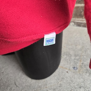 K&ouml;nnte beinhalten: Ein leuchtend rotes Sweatshirt mit einem kleinen wei&szlig;en Stoffetikett, das in den unteren Saum eingen&auml;ht ist. Das Etikett zeigt den Text Discus Athletic in blauen Buchstaben, eingerahmt von einem d&uuml;nnen gr&uuml;nen Rechteck auf wei&szlig;em Grund.