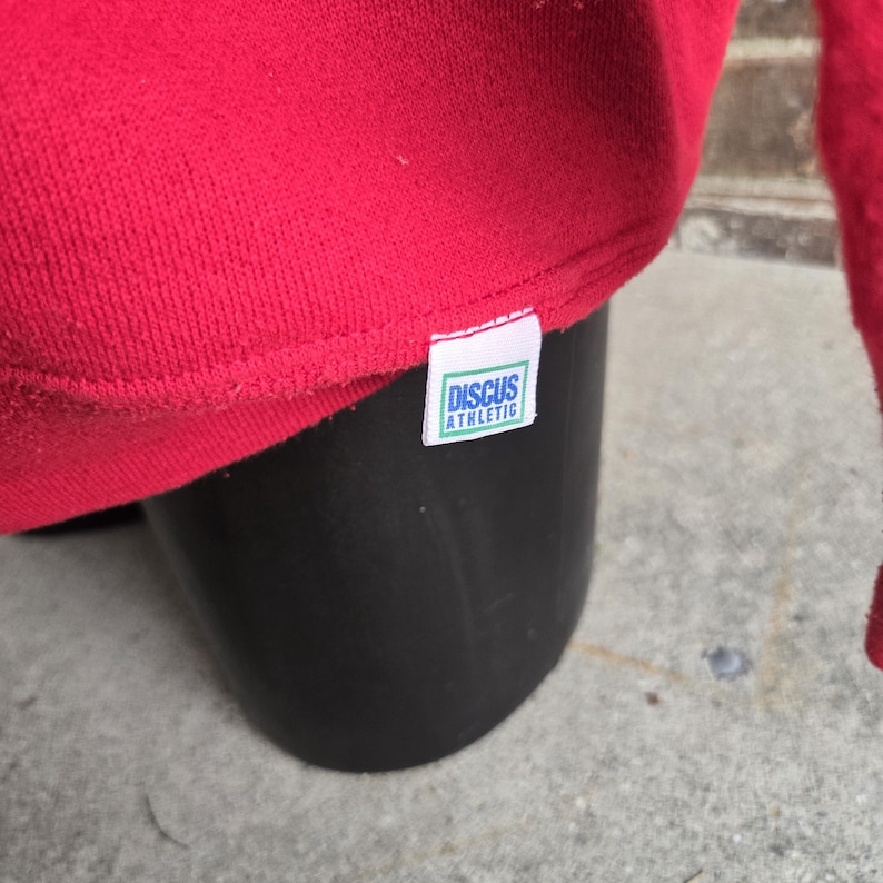 K&ouml;nnte beinhalten: Ein leuchtend rotes Sweatshirt mit einem kleinen wei&szlig;en Stoffetikett, das in den unteren Saum eingen&auml;ht ist. Das Etikett zeigt den Text Discus Athletic in blauen Buchstaben, eingerahmt von einem d&uuml;nnen gr&uuml;nen Rechteck auf wei&szlig;em Grund.