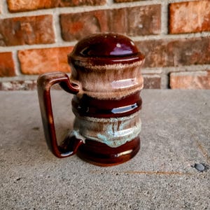May include: A ceramic salt or pepper shaker featuring a cylindrical body with a handle and a domed lid. The glaze is a blend of deep brown, creamy beige, and subtle teal tones, applied in horizontal bands with a glossy, smooth finish throughout.