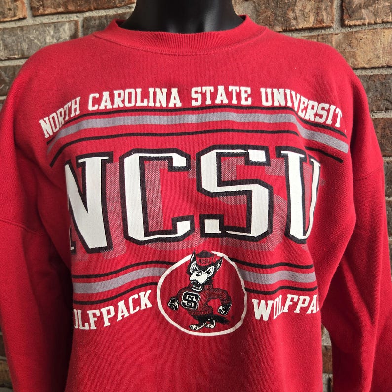 K&ouml;nnte beinhalten: Ein rotes Sweatshirt mit Rundhalsausschnitt, das oben den wei&szlig;en Schriftzug NORTH CAROLINA STATE UNIVERSITY und in der Mitte gro&szlig; NCSU zeigt. Darunter flankiert das Wort WOLFPACK eine kreisf&ouml;rmige Grafik eines Wolfsmaskottchens im roten Pullover.