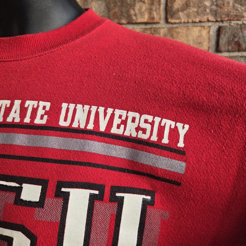 K&ouml;nnte beinhalten: Ein rotes Sweatshirt mit Rundhalsausschnitt und dem Schriftzug STATE UNIVERSITY in wei&szlig;en Gro&szlig;buchstaben. Unter dem Text befinden sich zwei horizontale Streifen in Grau und Schwarz, gefolgt von gro&szlig;en wei&szlig;en Buchstaben eines Logos.