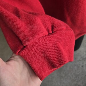 K&ouml;nnte beinhalten: Ein leuchtend rotes Langarm-Sweatshirt mit einem gerippten Strickb&uuml;ndchen am &Auml;rmelende. Der Stoff weist eine sichtbare, strukturierte Webart auf und der &Auml;rmel ist dort, wo er auf das elastische B&uuml;ndchen trifft, leicht gerafft.