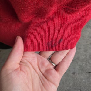 K&ouml;nnte beinhalten: Ein leuchtend rotes Rippstrickb&uuml;ndchen eines Kleidungsst&uuml;cks mit einem sichtbaren dunklen Fleck auf der unteren rechten Seite des Stoffes. Das Material hat eine texturierte, gewebte Optik mit einer durchgehend einheitlichen roten Farbe.