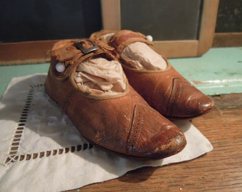May include: A pair of brown leather baby shoes with a buckle closure and white pearl accents. The shoes are worn and have a vintage look.