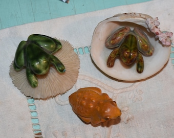 May include: Three ceramic frog figurines. One green frog is sitting on a white coral-like base. Another green frog is sitting inside a white seashell. The third frog is brown and sitting on a white surface.