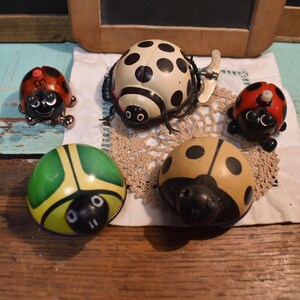 May include: A collection of vintage ladybug toys in various colors and designs. The ladybugs are red, yellow, green, and cream with black spots. Some have wind-up mechanisms and detailed features, arranged on a lace doily.