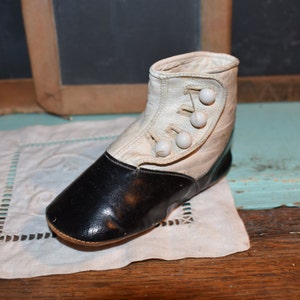 May include: Vintage baby bootie in black and white leather. The boot has a rounded toe and three white buttons on the side. The boot is sitting on a white embroidered cloth.