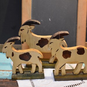 May include: Three wooden goat figurines. The goats are painted with a light tan base color and dark brown spots. The horns and bases are also painted brown. The goats are standing on green bases.