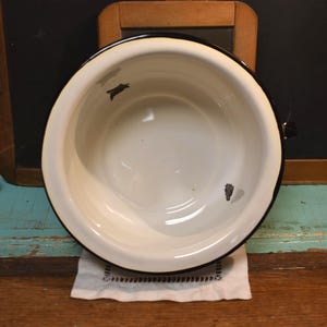 May include: An overhead view of a white enamelware bowl with a black rim and handle. The bowl has some wear and tear, with visible dark spots on the interior. The bowl sits on a white cloth with a decorative edge.