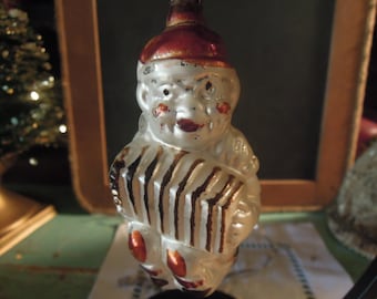 May include: Vintage Christmas ornament shaped like a musician playing an accordion. The ornament has a white body with brown stripes on the accordion, a red hat, and red accents on the feet. The ornament is made of glass.