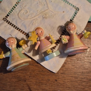 Może przedstawiać: Trzy figurki drewnianych aniołów w stylu vintage z malowanymi detalami. Każdy anioł ma unikalny design, trzymając przedmioty takie jak gwiazda, dzwonek i prezent. Anioły znajdują się na białej, haftowanej tkaninie, na drewnianej powierzchni.