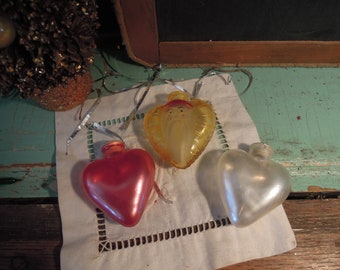 May include: Three vintage Christmas ornaments in heart shapes. One is red, one is pearlescent white, and the third is yellow with a Santa Claus face. They are on a white cloth with a decorative border.
