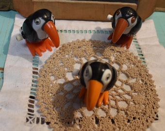 May include: Three vintage wind-up penguin toys. The penguins have black bodies, white faces, orange beaks and feet. They are arranged on a beige crocheted doily. The background includes a wooden chalkboard and a light blue surface.